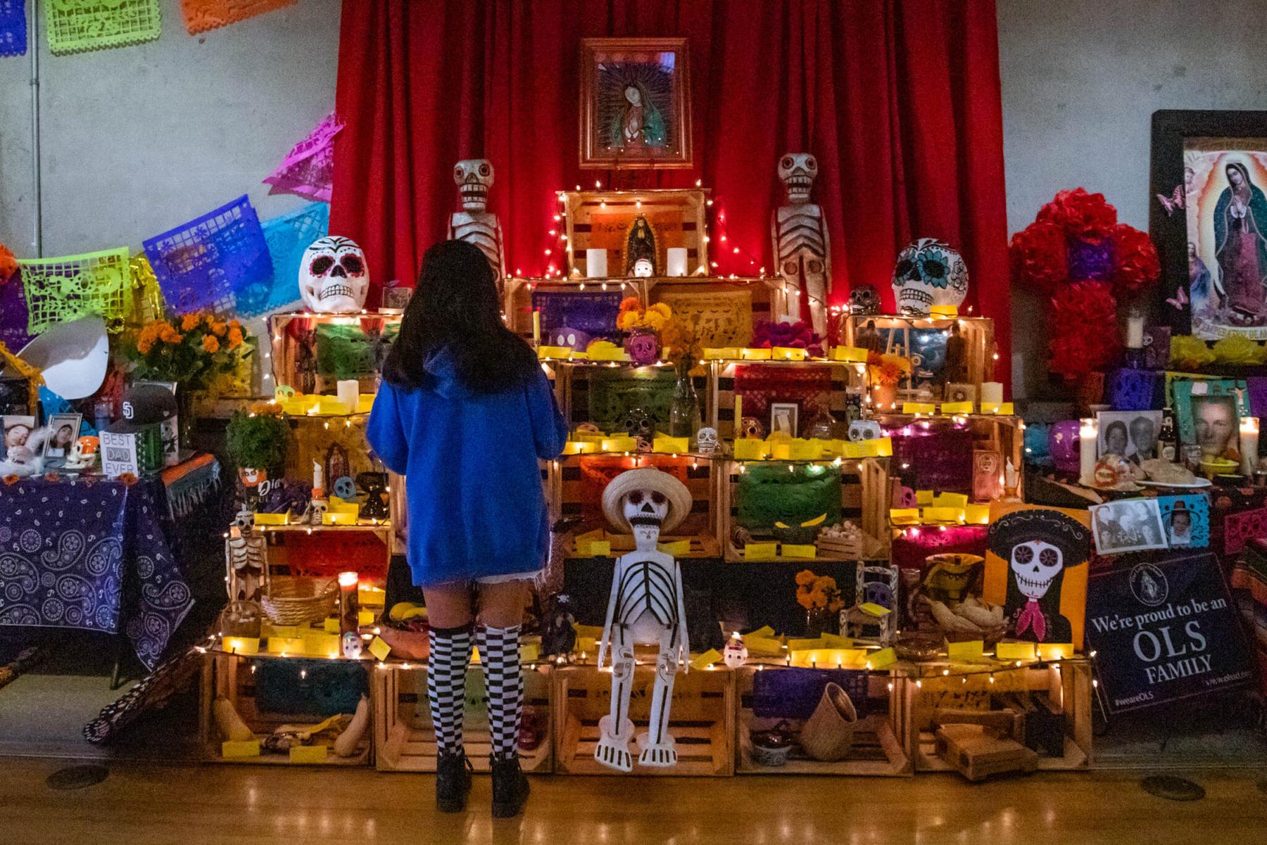 A visitor standing before a towering community ofrenda of stacked wooden crates, sugar skulls, marigolds, candles and family photographs