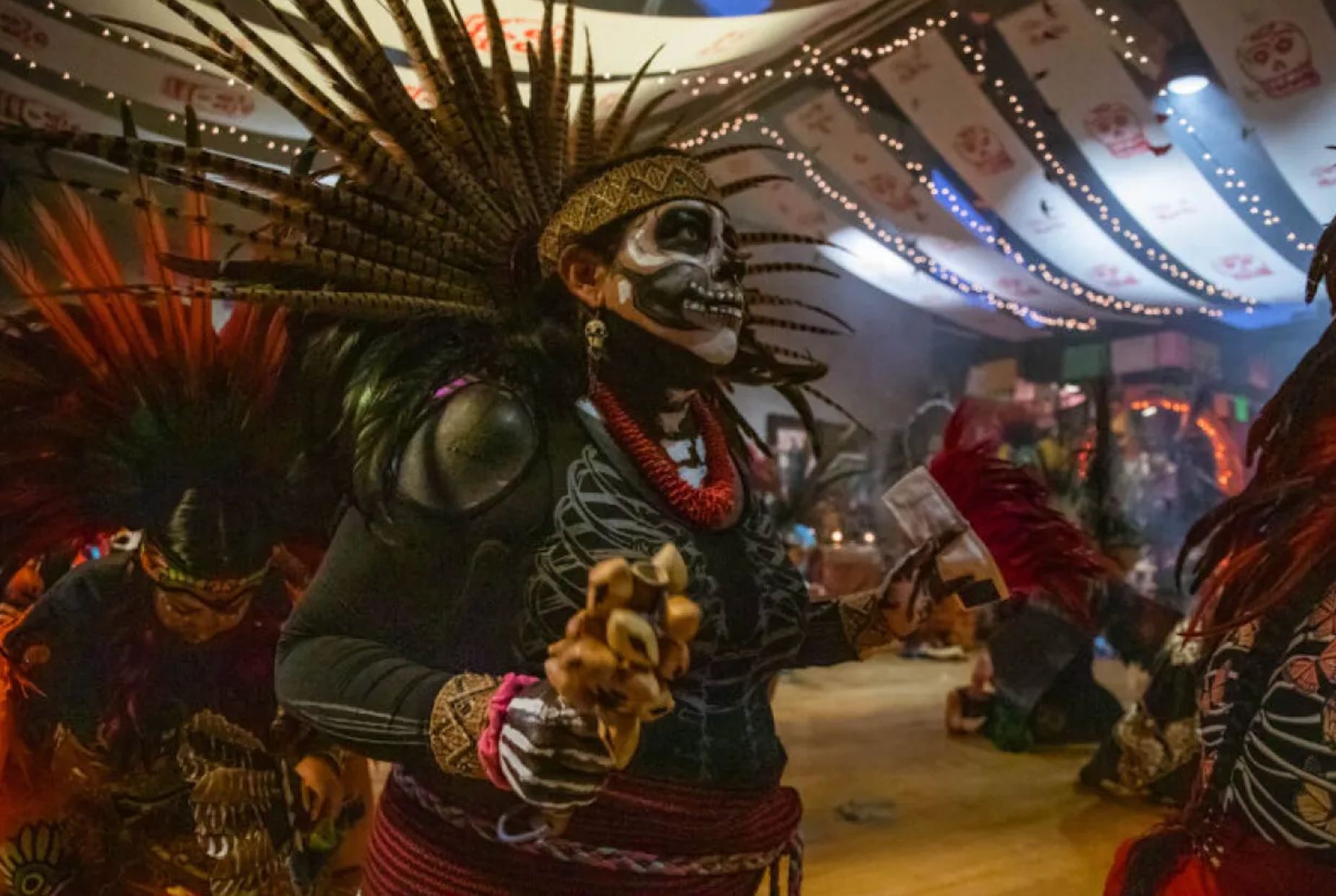 Danzante in a feathered headdress and calavera face paint, mid-dance inside the Center beneath strings of lights and papel picado