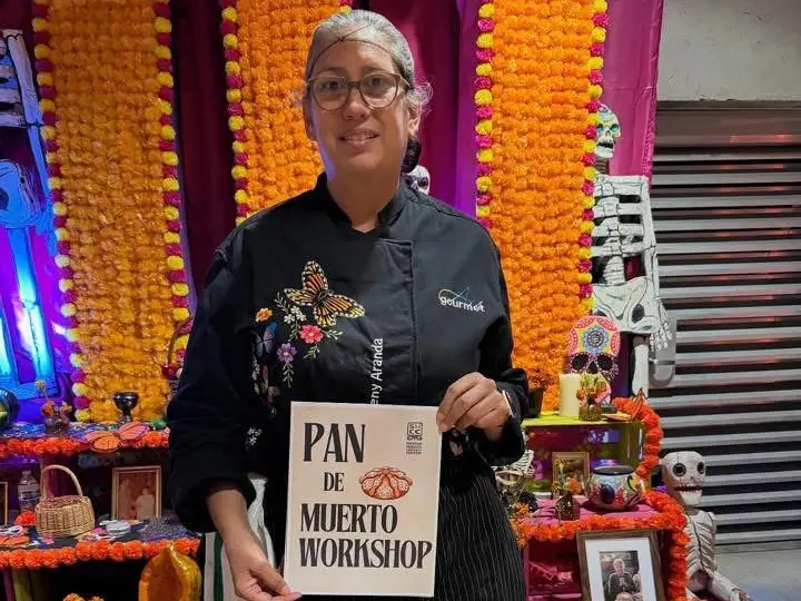 Chef Aureny Aranda holding a Pan de Muerto Workshop sign in front of a Día de los Muertos altar